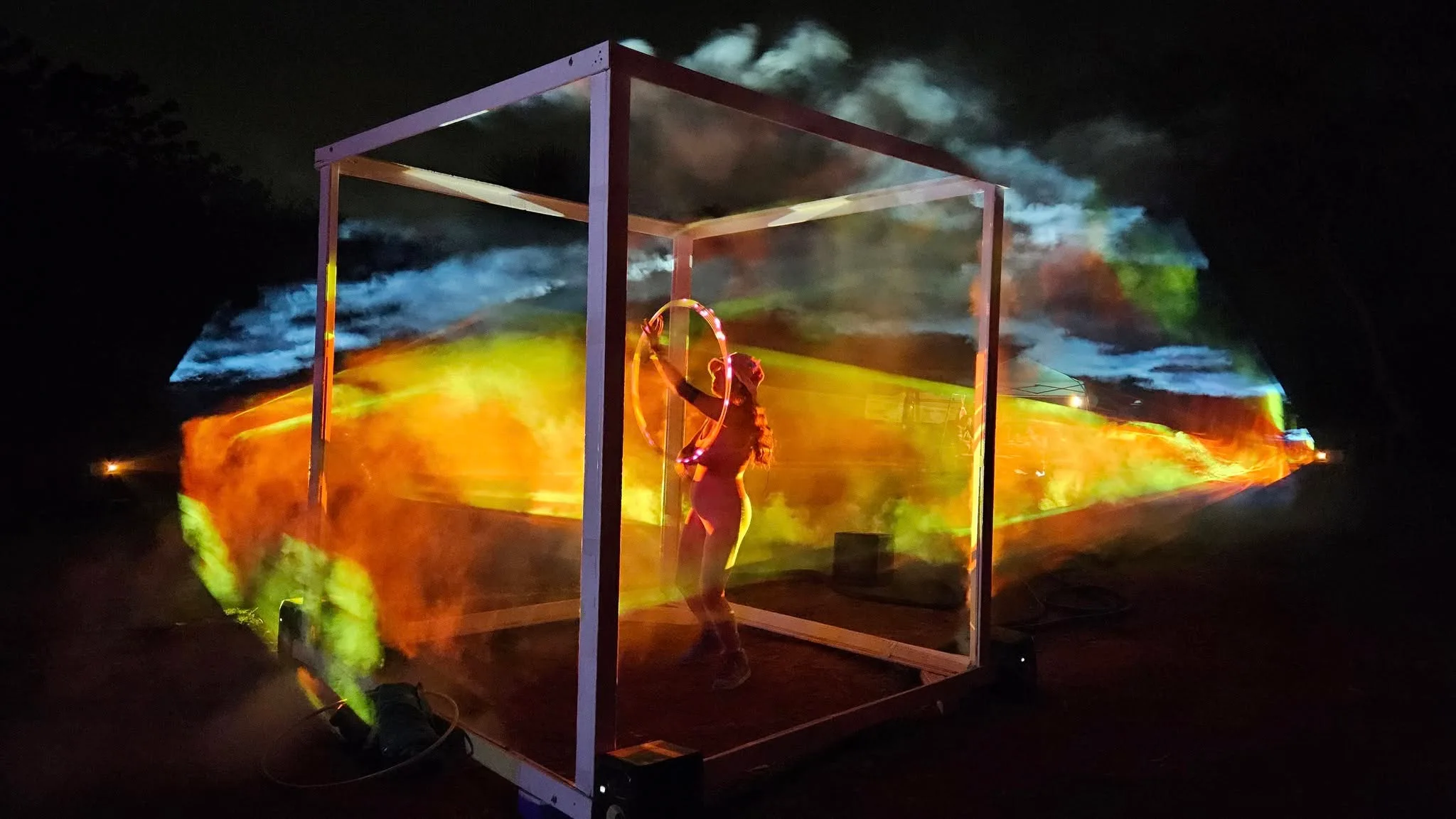 At night, a glass cube glows with orange and blue holographic clouds while a woman inside holds a lit hoop above her head.