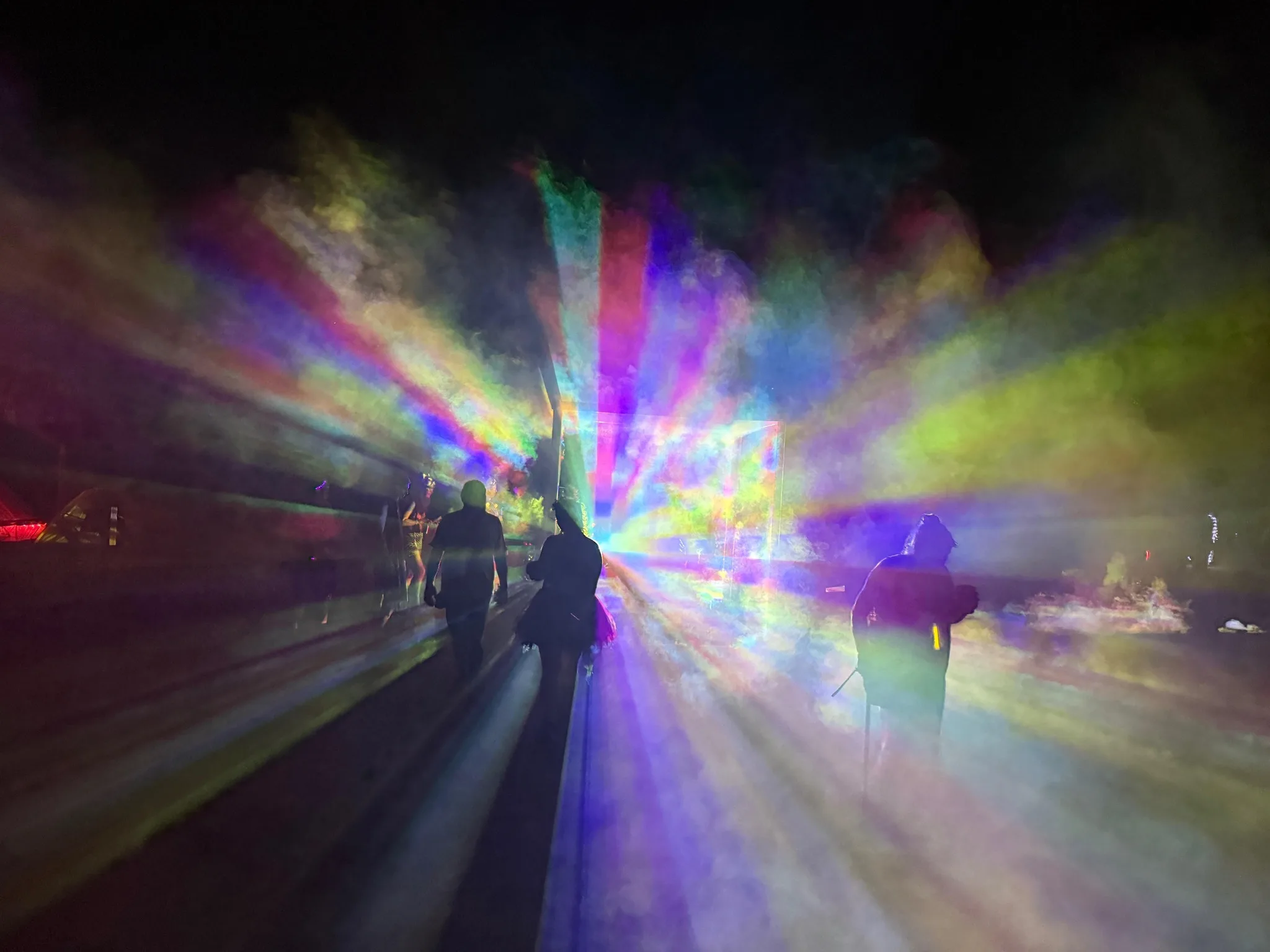 Rainbow-colored light rays radiate through thick fog as three silhouetted people walk past, with the glowing cube visible in the distance.