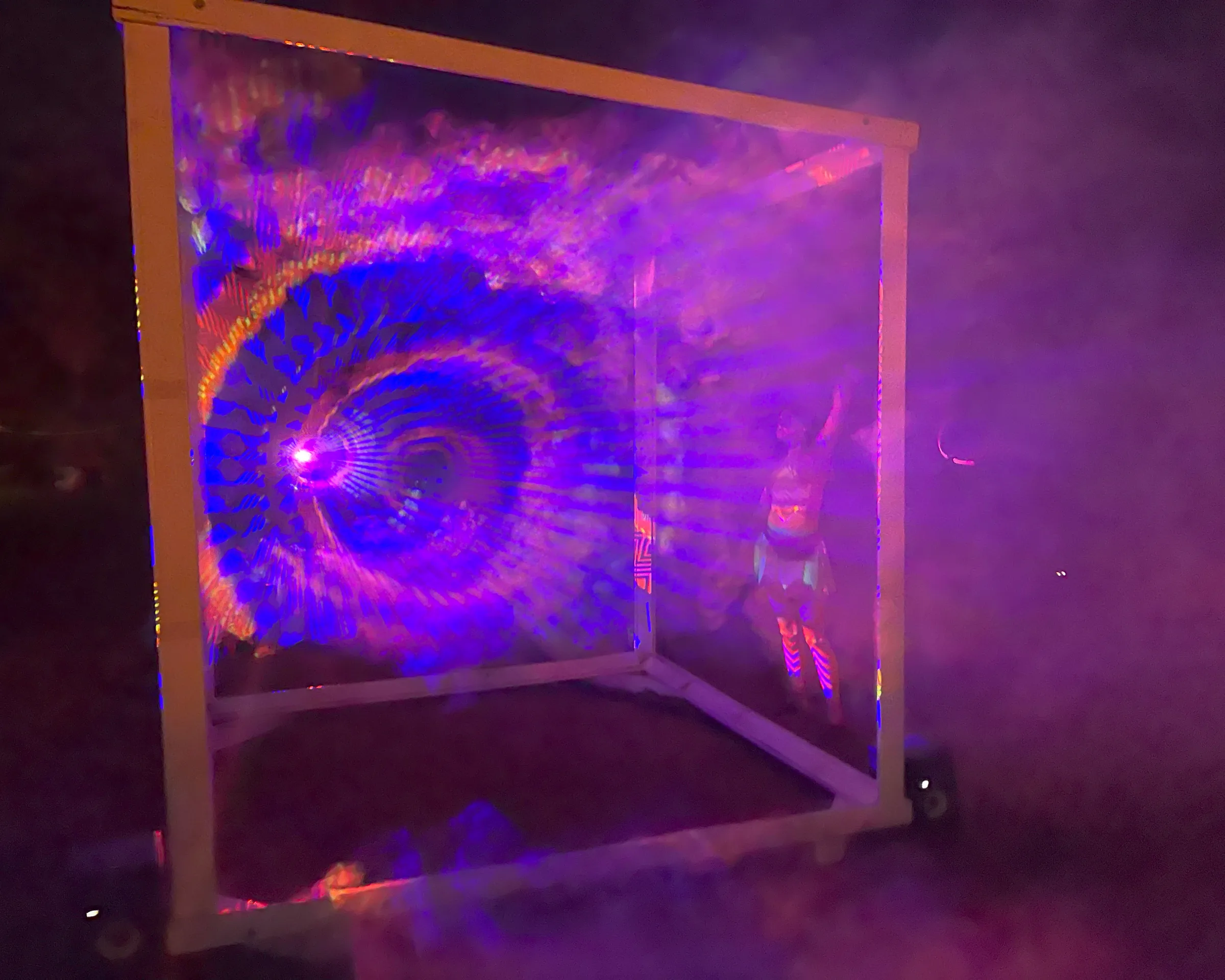 A swirling blue and purple spiral hologram fills one panel of the cube at night, with a person in striped leggings standing inside amid thick fog.