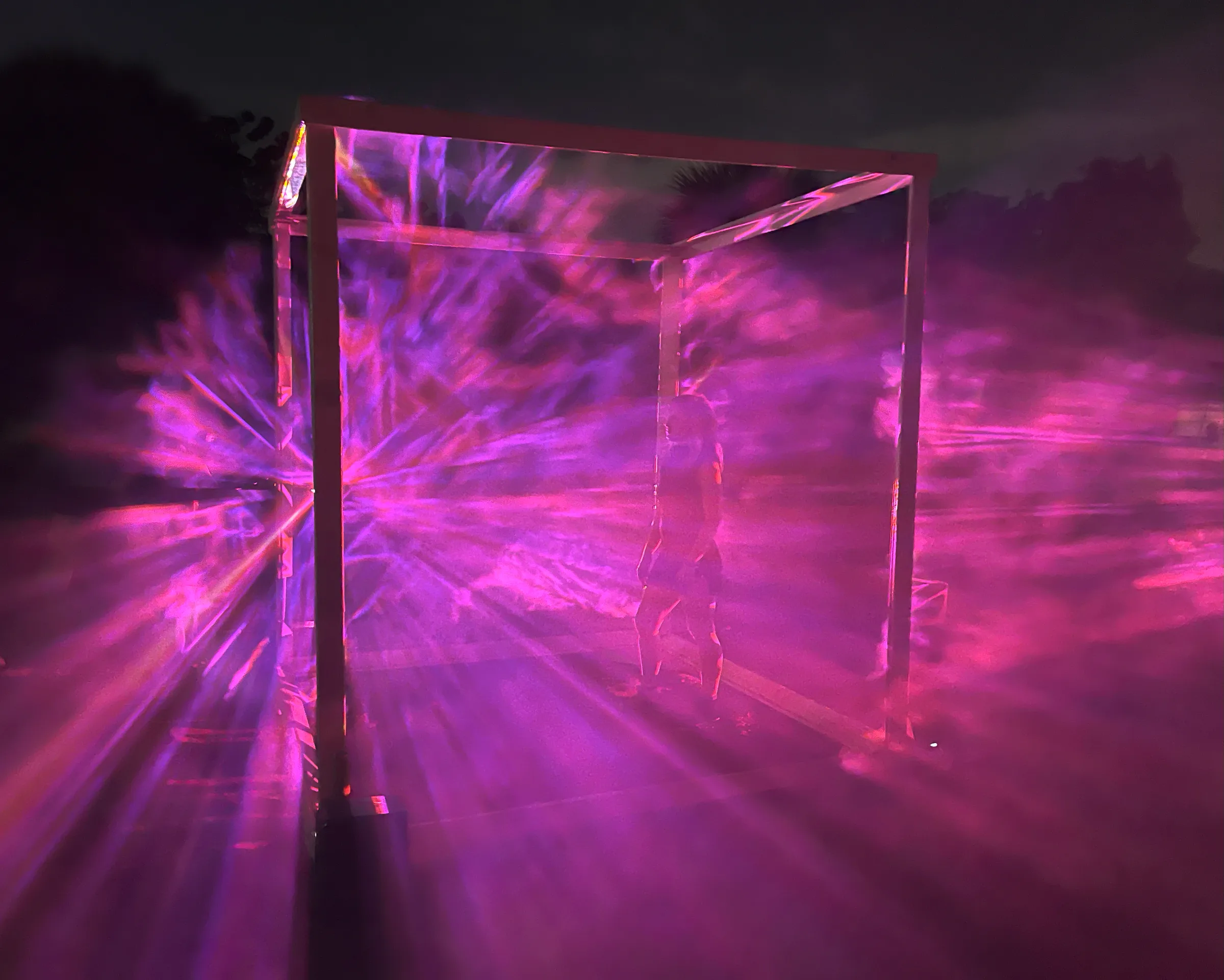 Magenta and purple light rays burst across the cube's panels at night, silhouetting a single person standing inside.