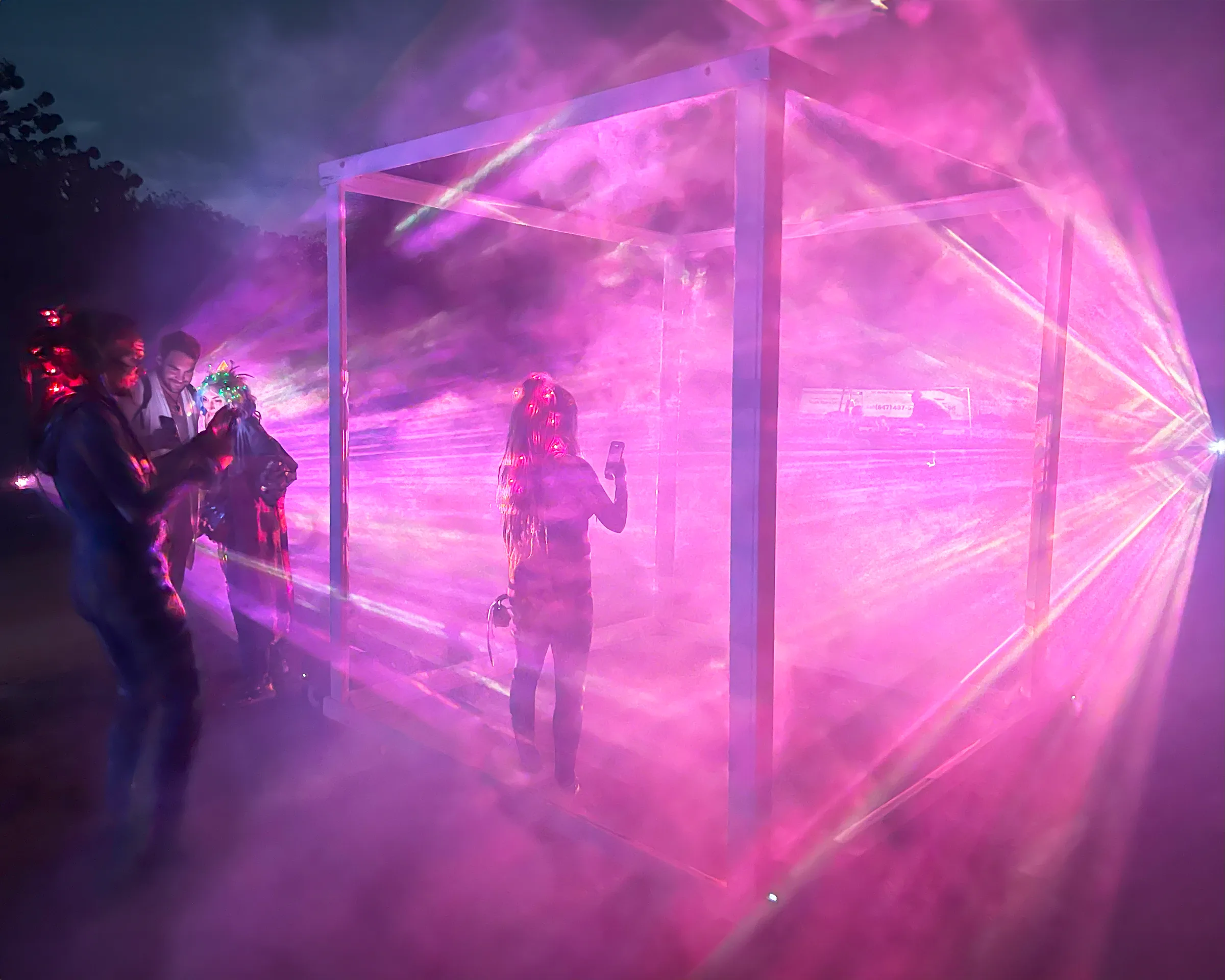 Pink fog glows inside the cube as a person films on their phone, while a small group of onlookers watches from just outside.