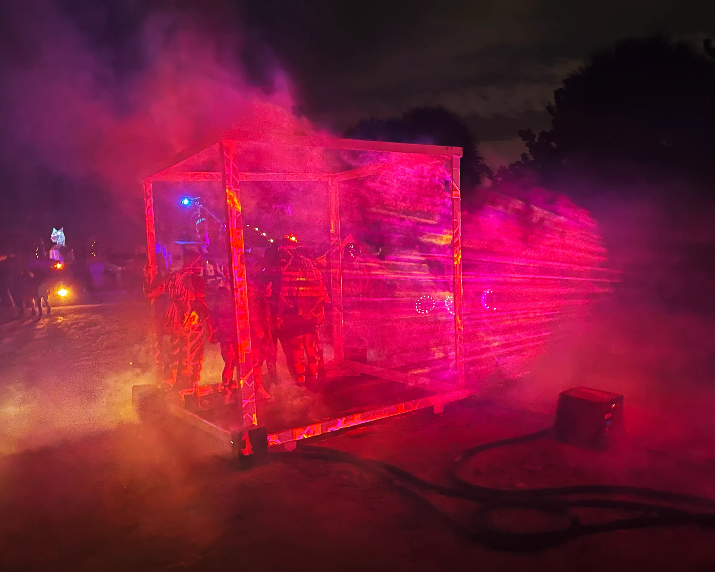 Deep red and magenta fog engulfs the cube as several people in patterned costumes gather inside, with glowing ring props and a crowd nearby.