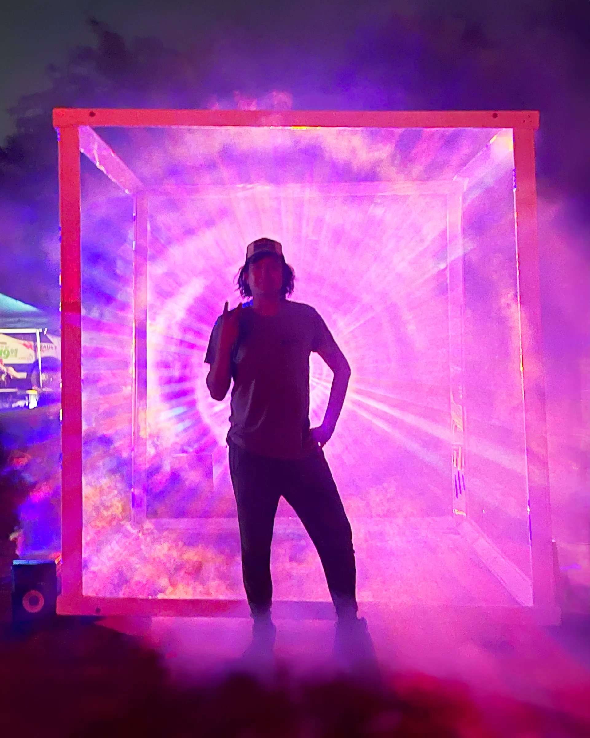 A person in a cap stands silhouetted inside the cube, backlit by radiating pink light and fog, with a truck and tent visible behind them.