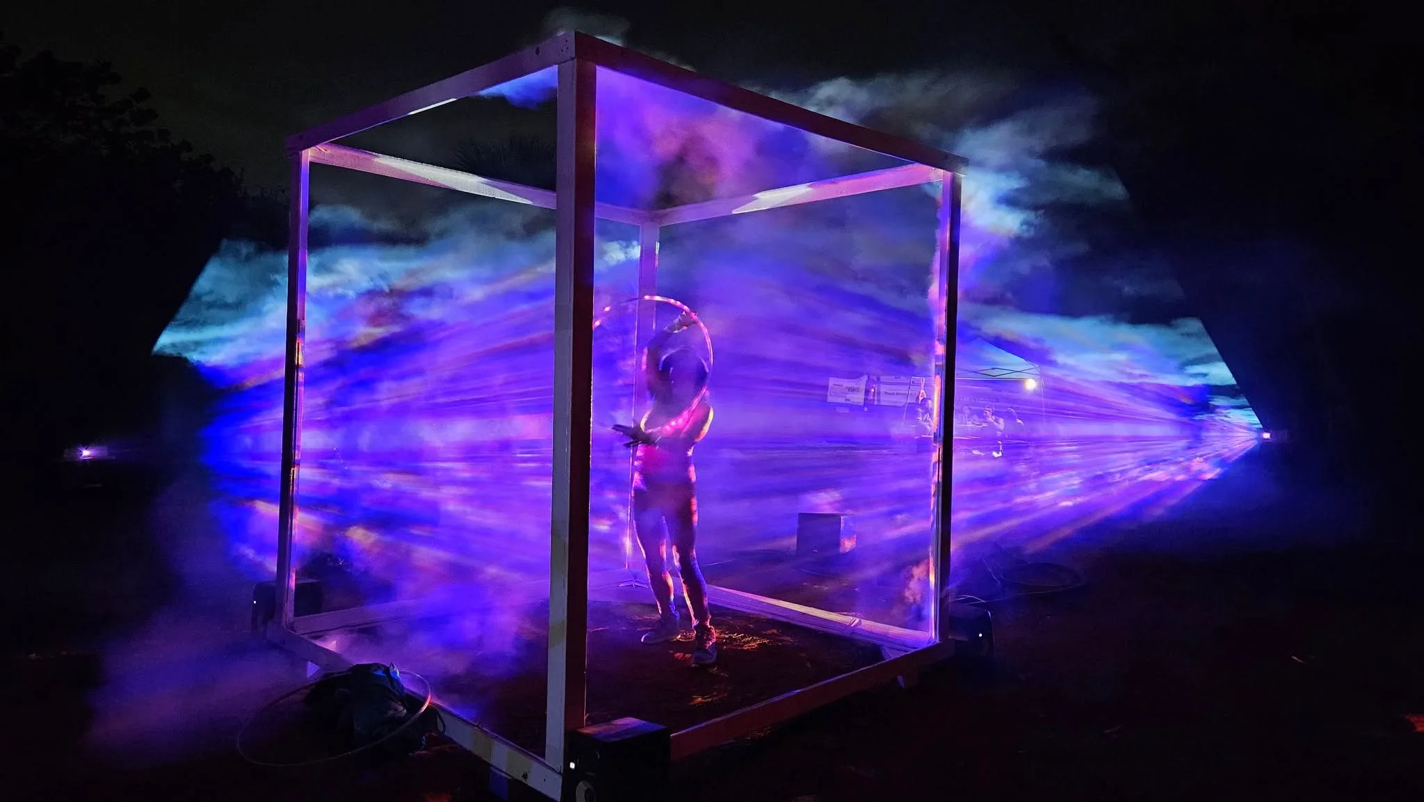 Blue and purple cloud-like holograms fill the cube at night while a person inside raises a glowing hoop overhead.