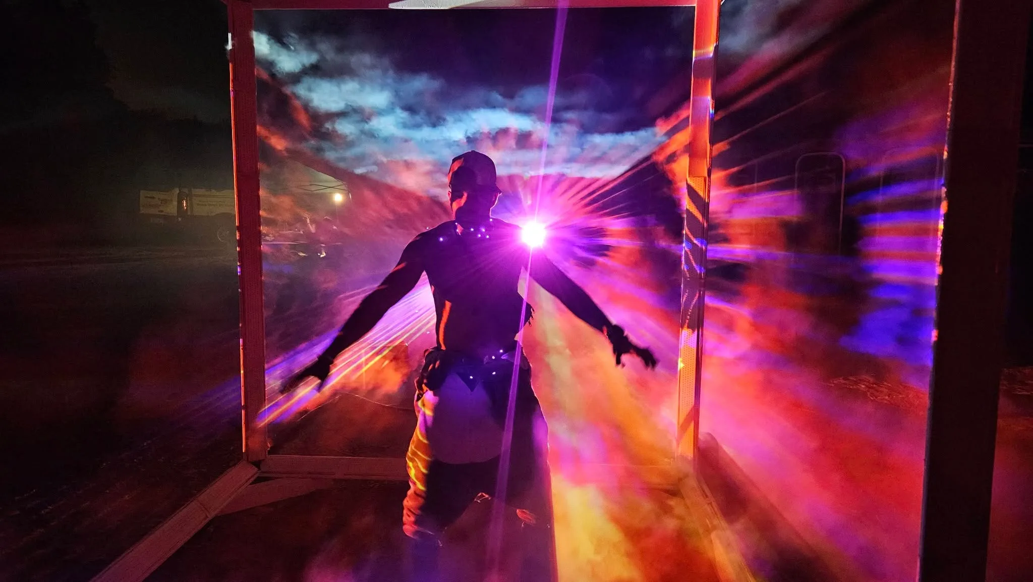 A silhouetted figure spreads their arms wide inside the cube, backlit by a bright pink light against orange and purple projected clouds.