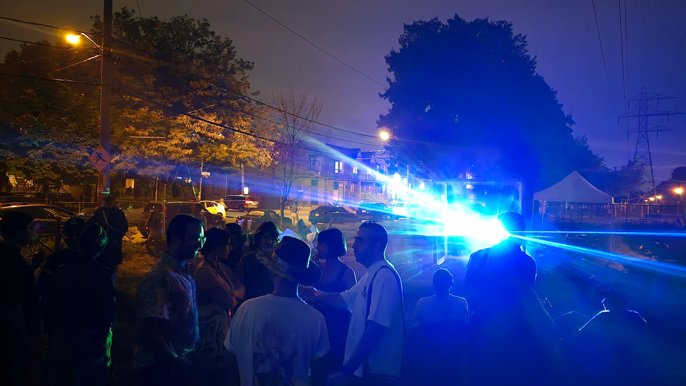A crowd gathers on a street at dusk as intense blue laser beams shoot from the distant cube installation past houses and power lines.