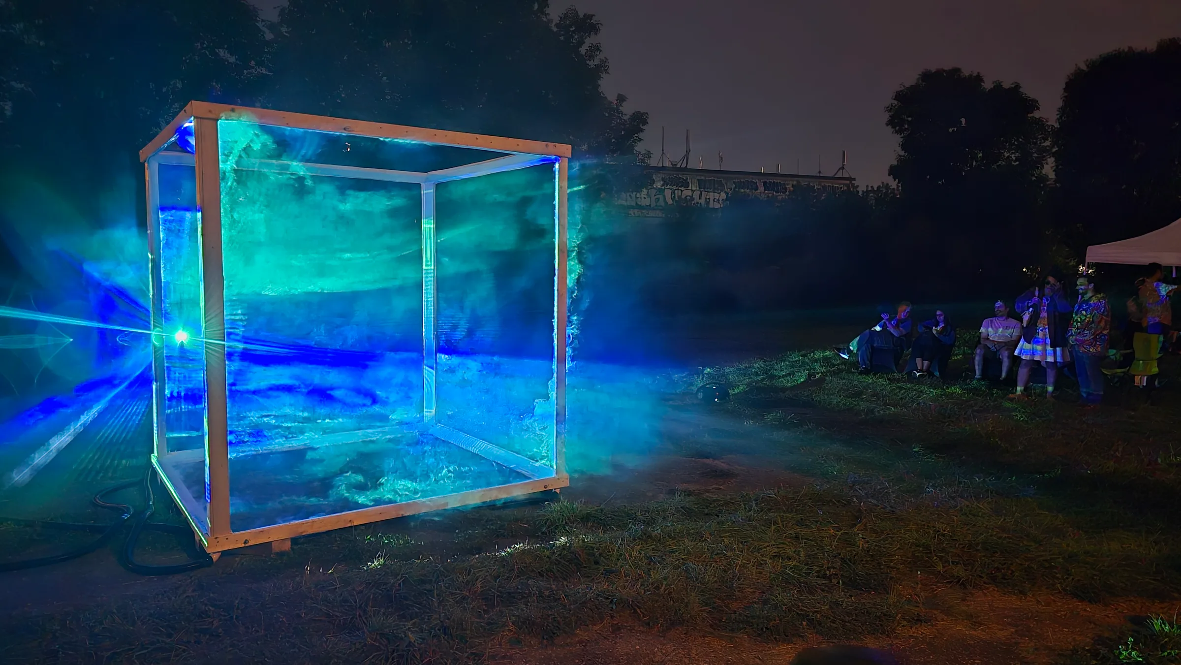 The cube glows teal and blue like a fog-filled tank in a grassy field, as costumed onlookers watch from a distance.