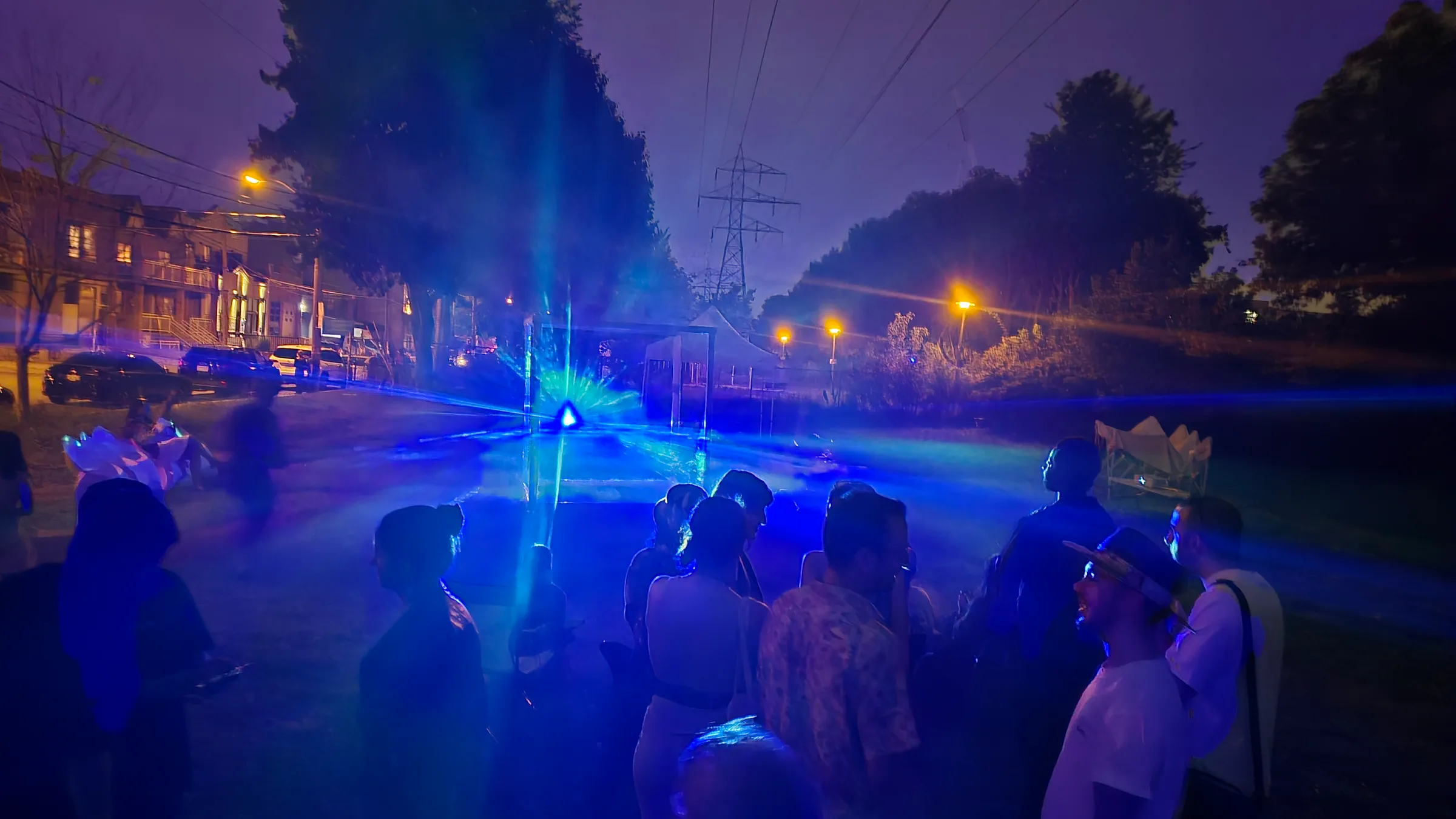 A dense crowd stands bathed in blue light as the distant cube emits a triangular beam above a residential street at night.
