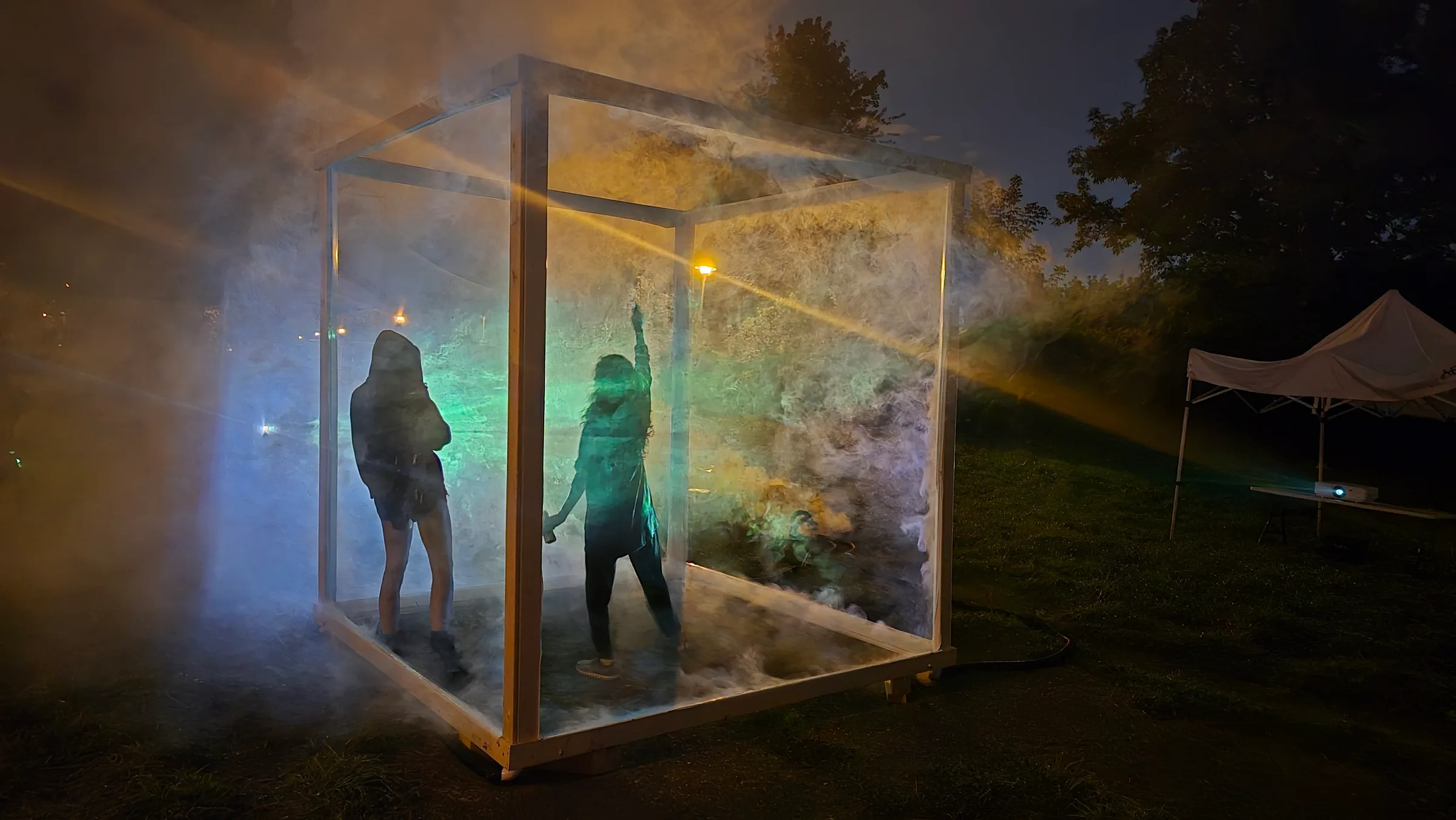 Two silhouetted people dance inside the fog-filled cube, lit in teal and green, one reaching an arm upward.