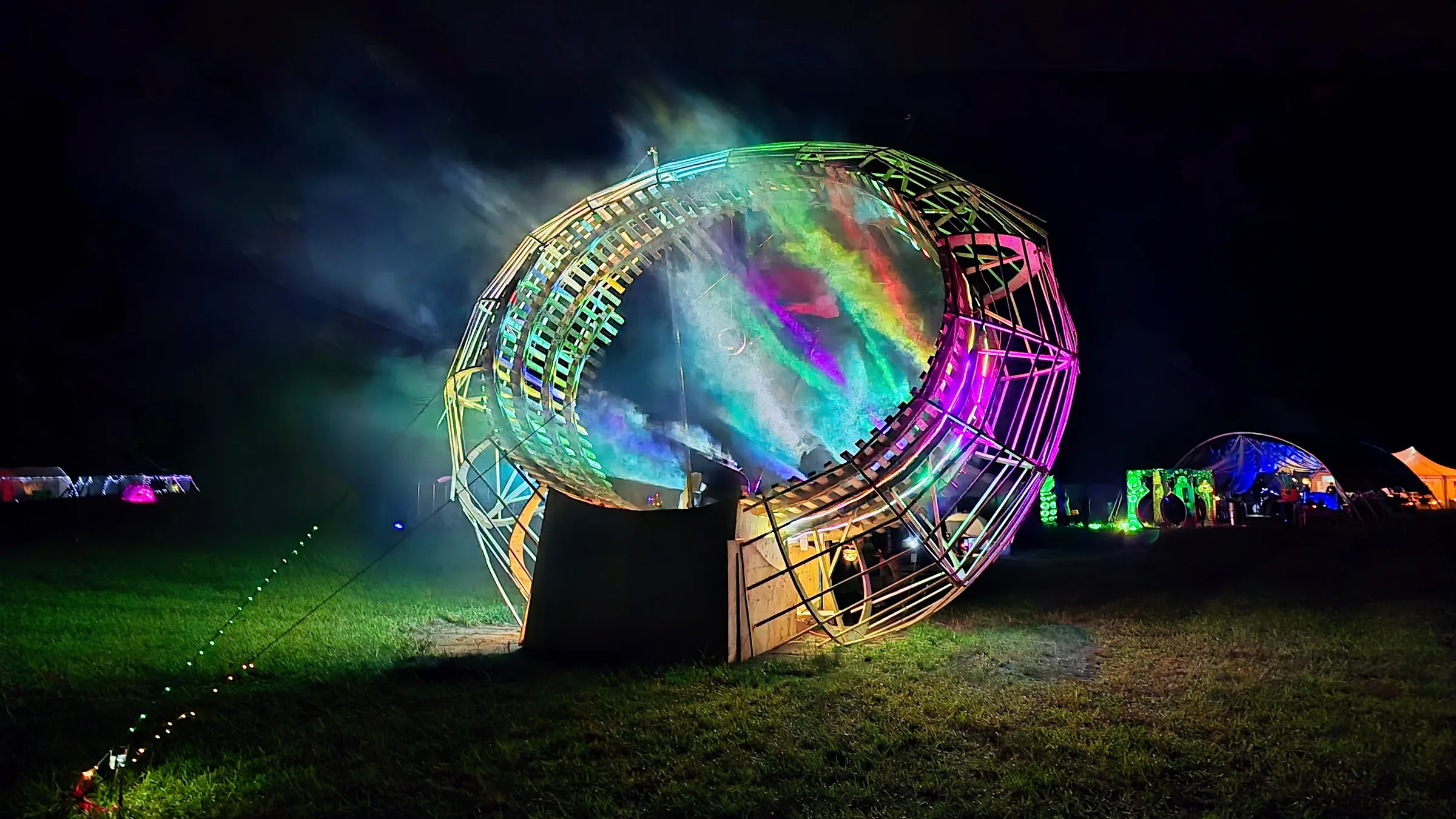 A large wooden ring glows green and magenta at night, its open center filled with rainbow holographic mist above a grassy festival field.