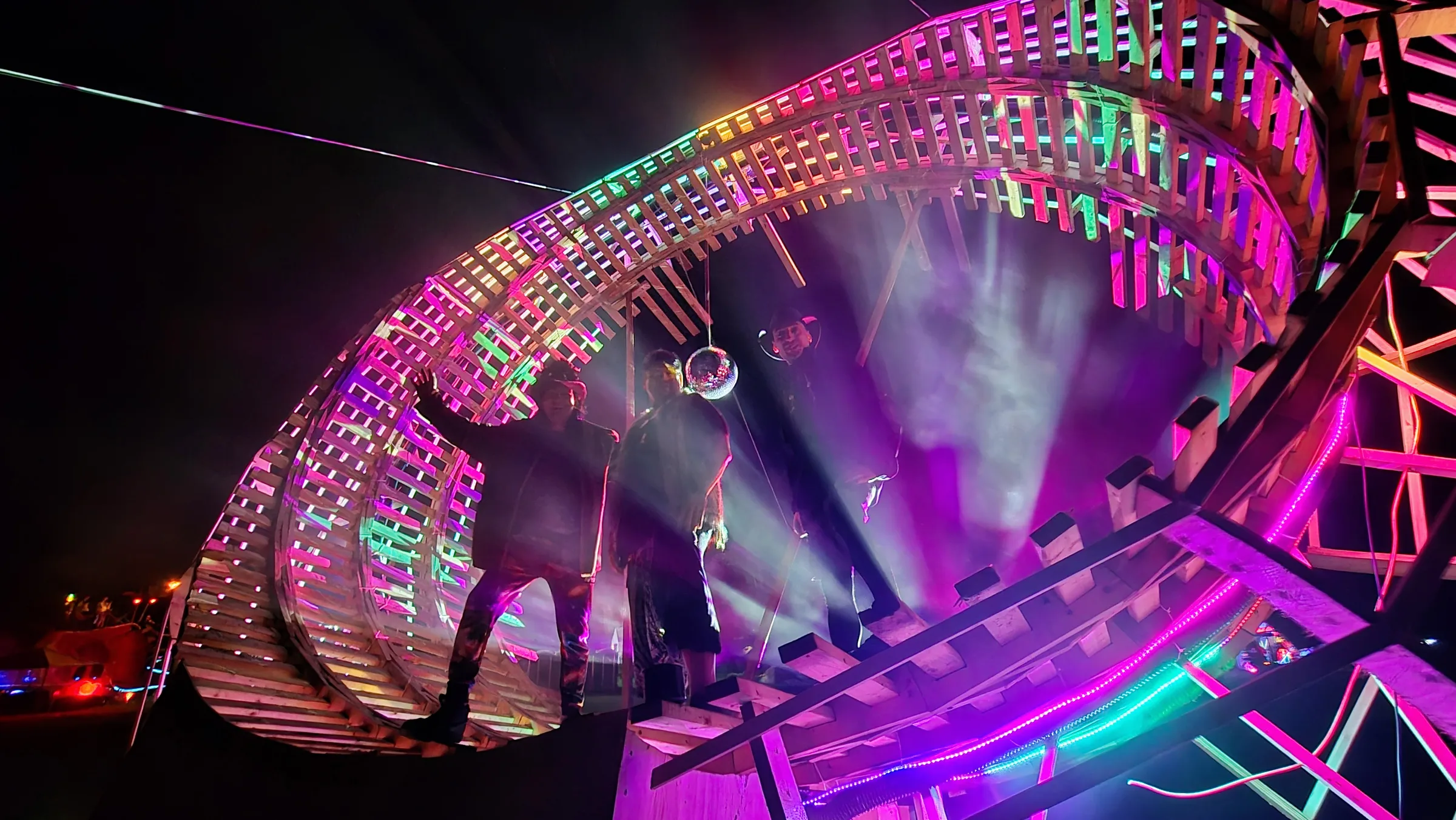 Three silhouetted people stand beneath the glowing wooden ring at night, purple and white light beams cutting through haze near a disco ball.