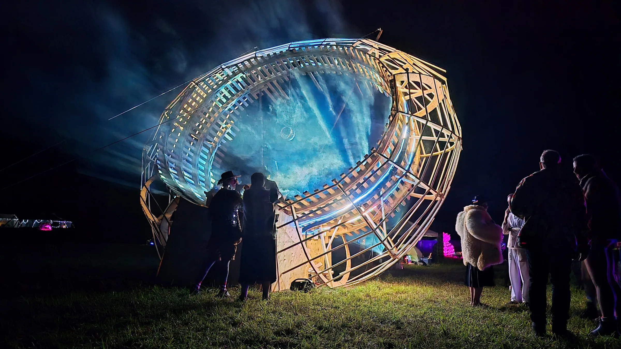 Spectators watch the wooden ring illuminated with blue and teal holographic mist at night, other festival lights glowing in the background.