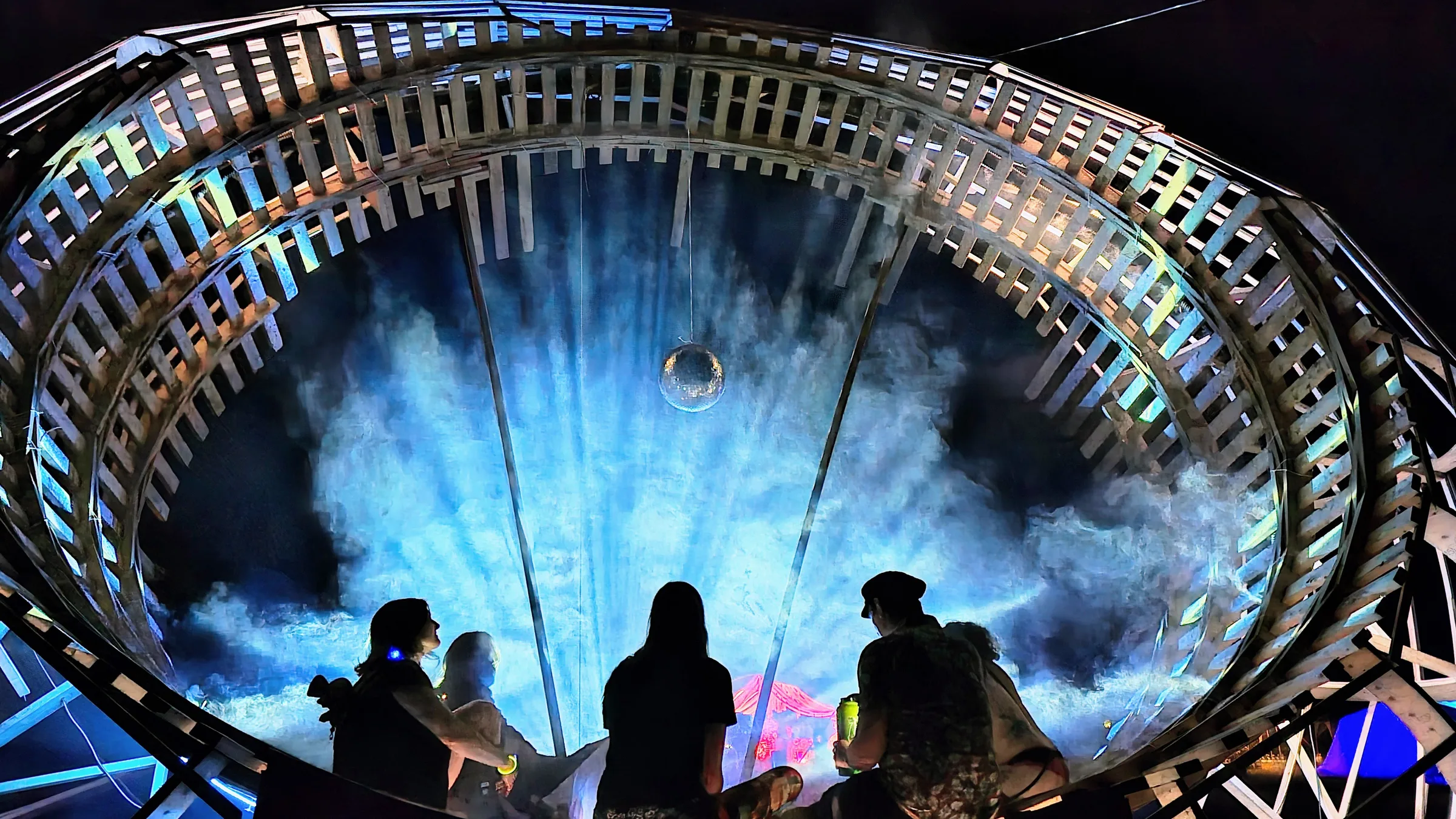 Viewed from below, three silhouetted festivalgoers look up into the wooden ring's glowing blue mist, a small disco ball hanging at its center.