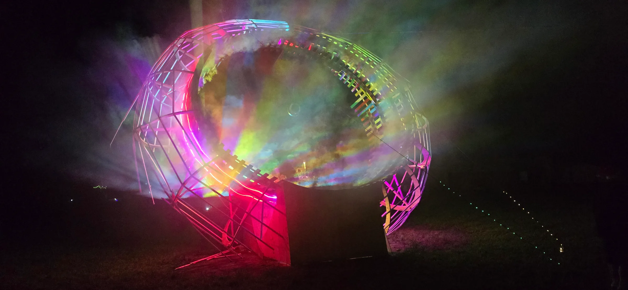 A wooden ring installation glows pink and green at night, filled with swirling holographic mist, a string of small lights trailing across dark grass.