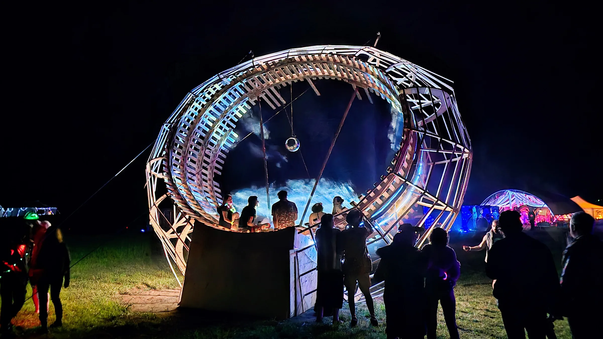 A crowd gathers on the grass to watch the wooden ring illuminated with blue mist at night, a disco ball hanging inside and colorful tents beyond.