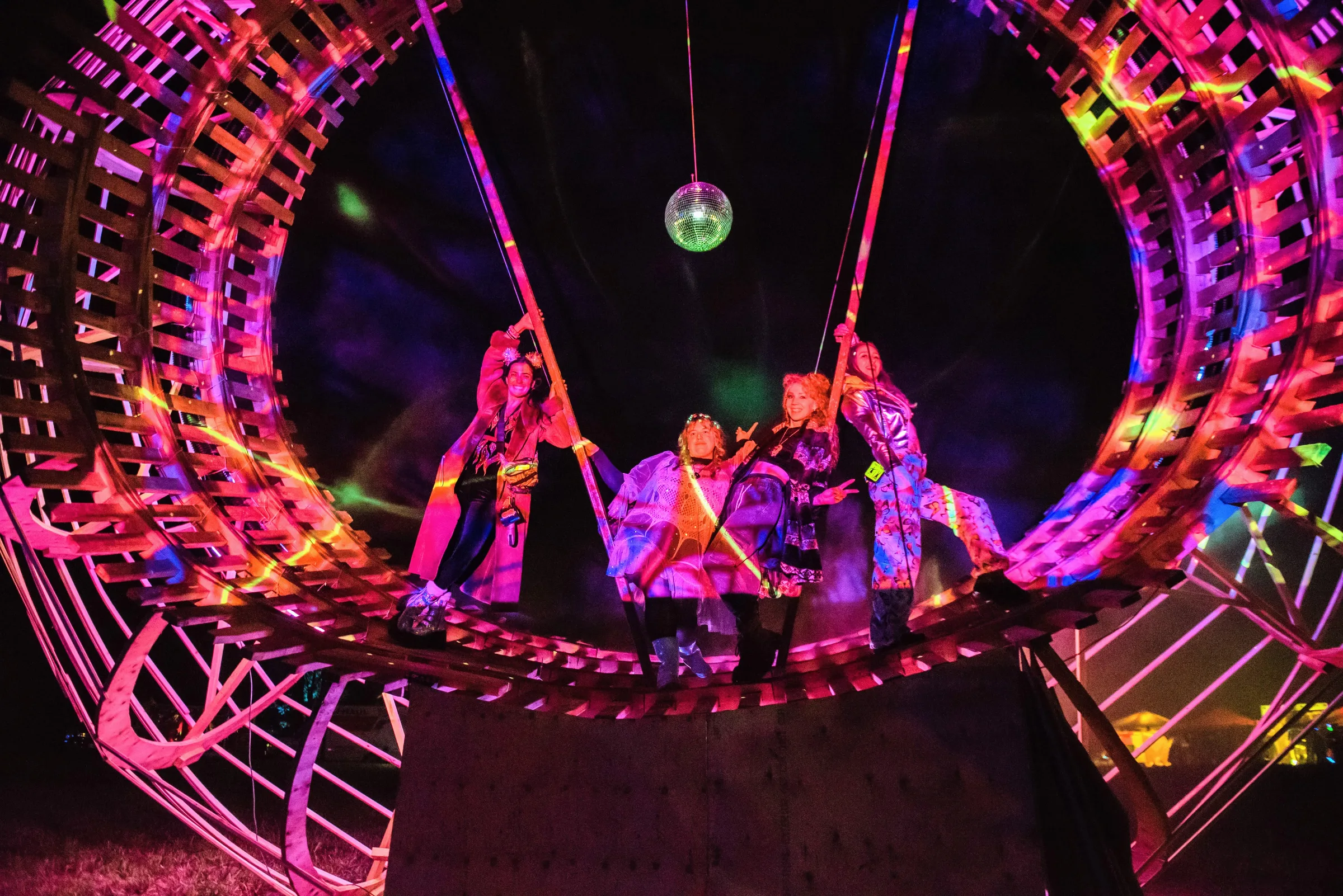Four festivalgoers pose on the ring's wooden platform lit in hot pink, holding overhead ropes beneath a hanging disco ball at night.