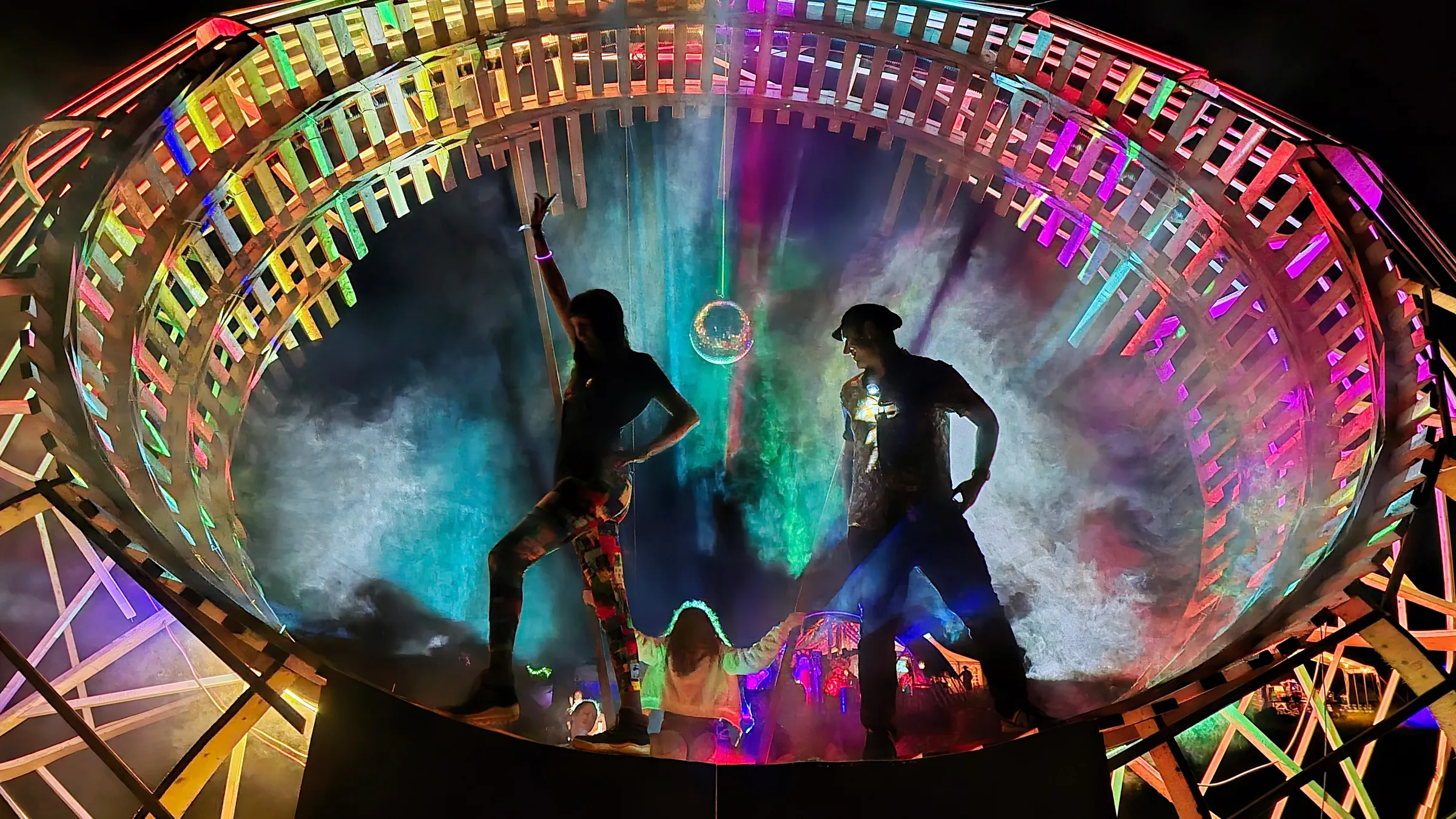 Two silhouetted figures strike poses on the ring's platform at night, a child between them, teal mist glowing behind a hanging disco ball.