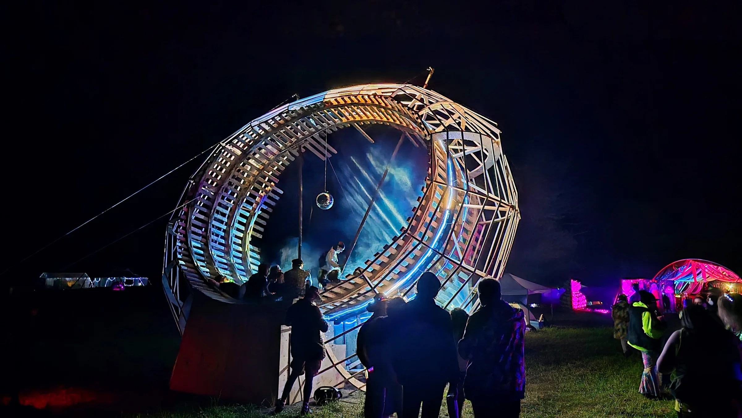 Spectators stand before the wooden ring glowing blue at night, a disco ball hanging inside and a pink lit tent visible to the side.