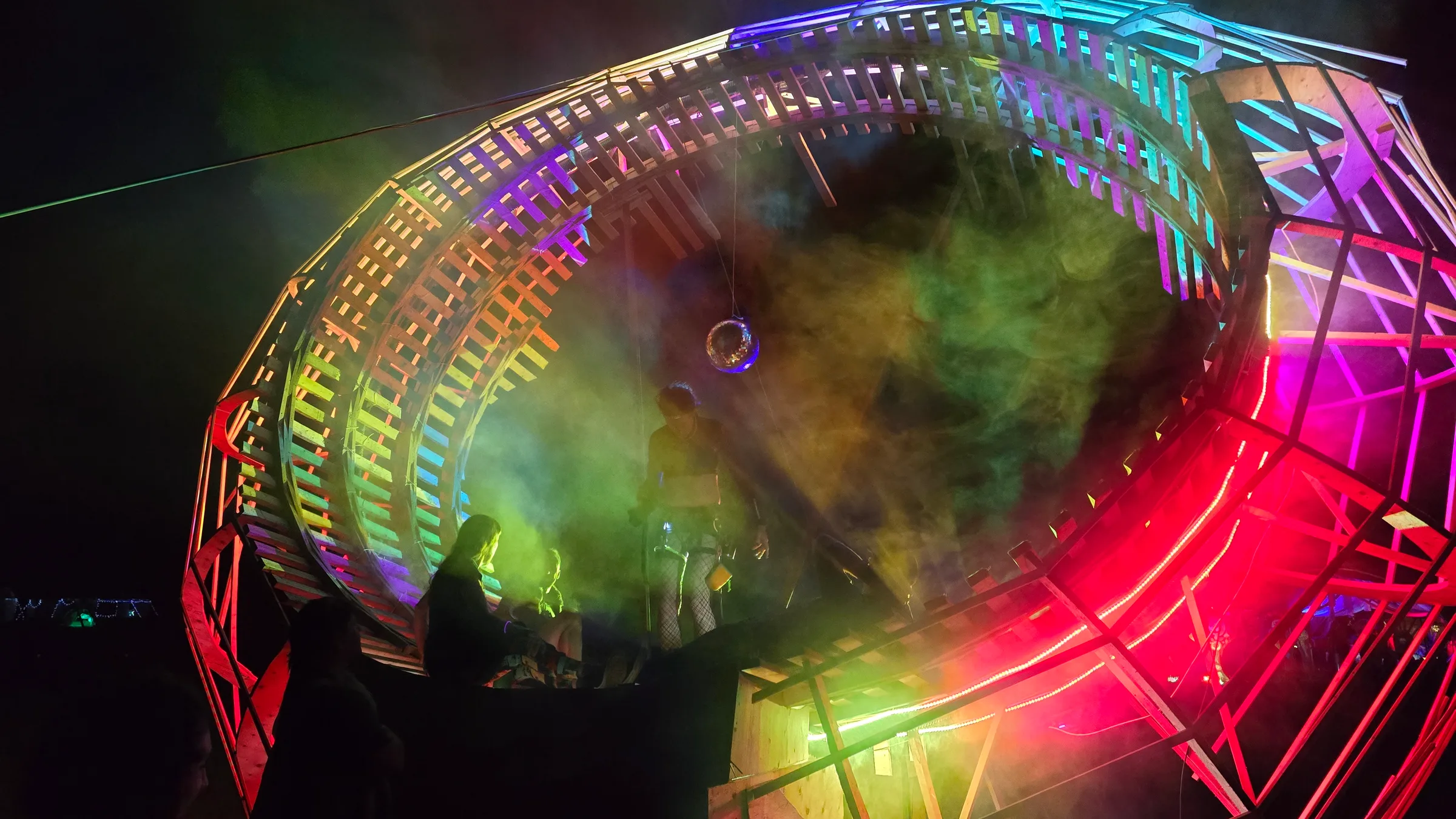 Looking up into the ring, hazy green mist fills the center around a disco ball, with silhouetted figures on the platform and red LED trim below.