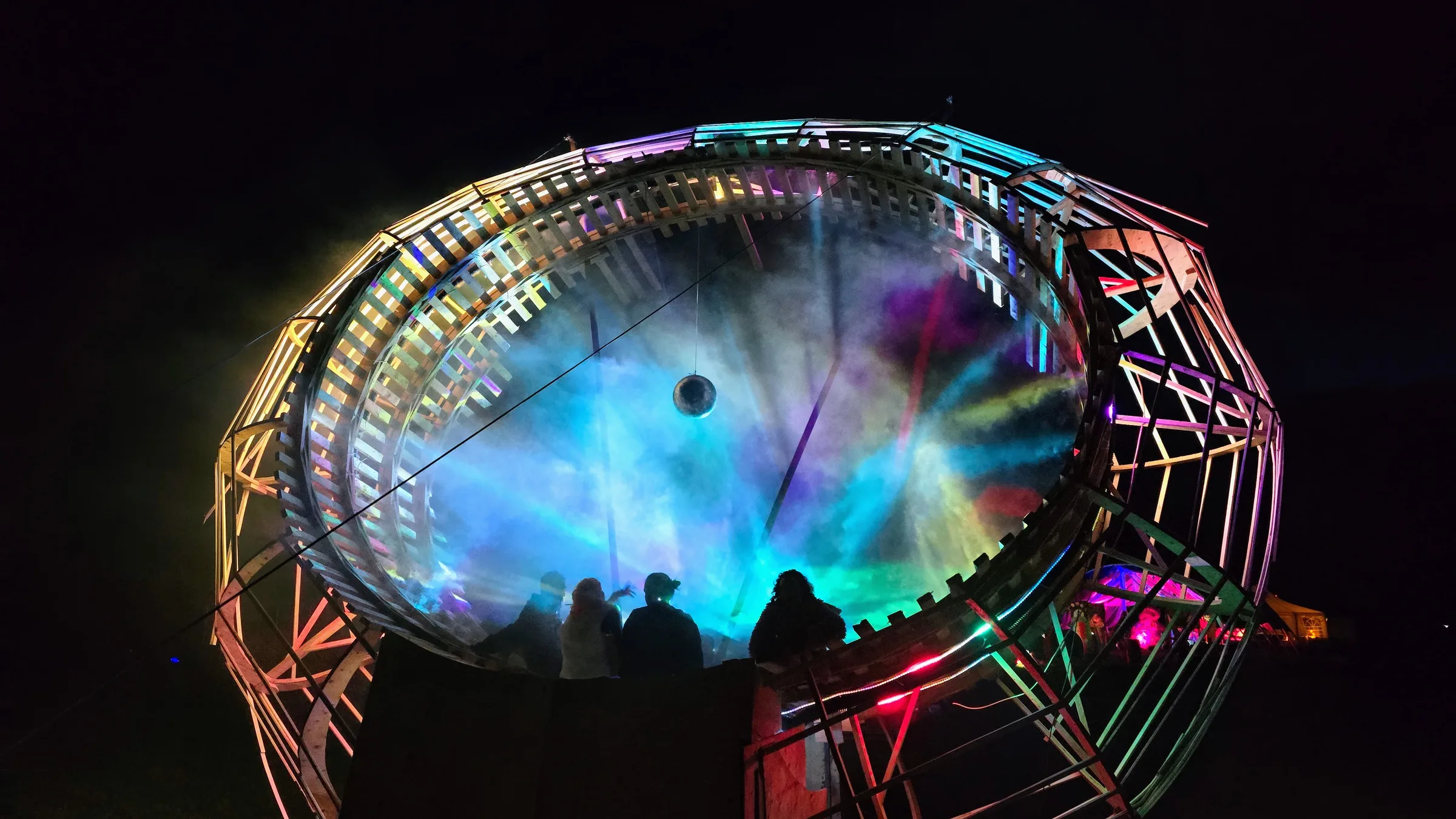 Rays of blue, purple and teal light fan through misty air inside the ring at night, silhouetted onlookers standing beneath a small disco ball.