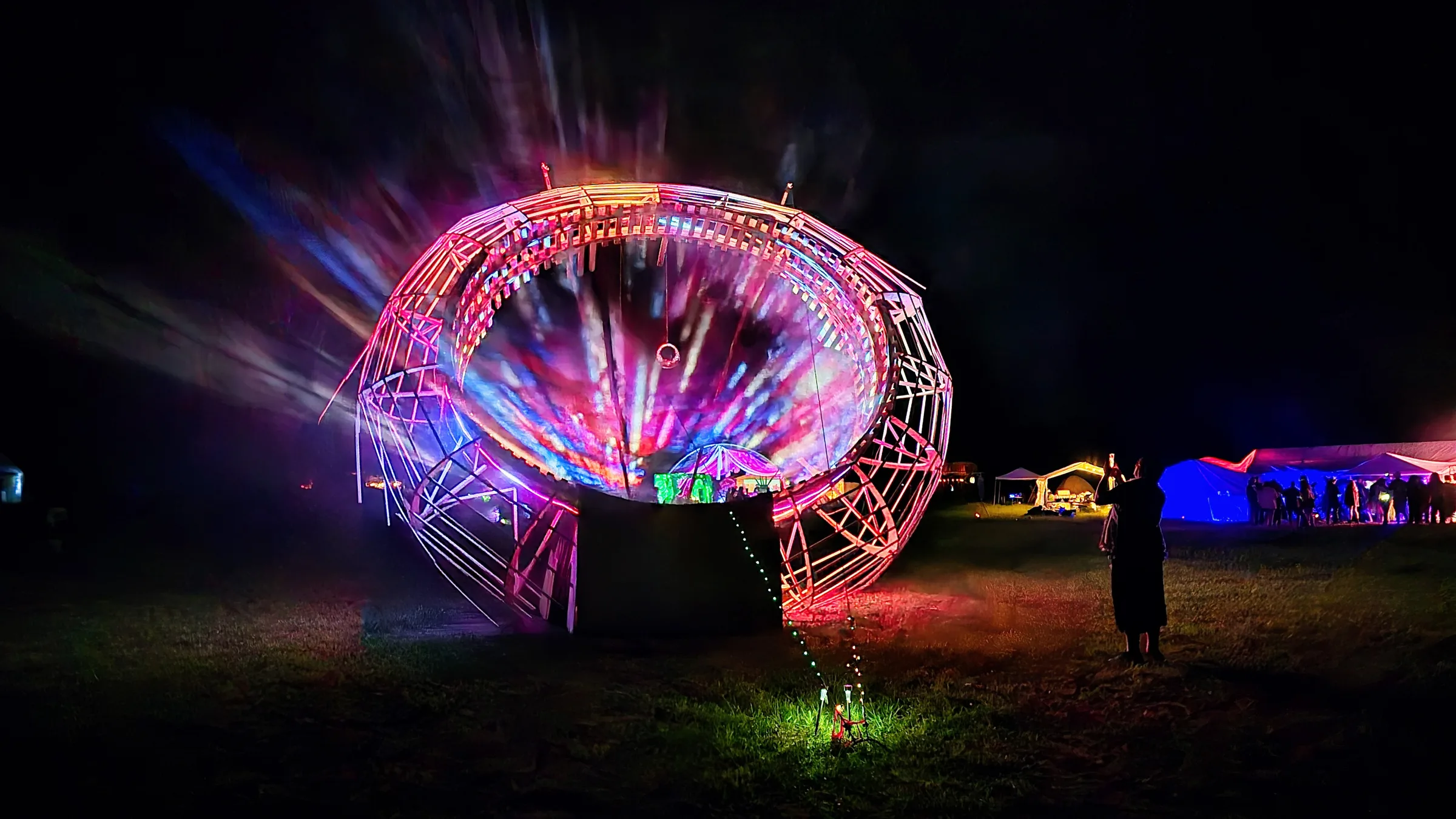 A wide view of the wooden ring at night, pink and blue light beams radiating outward as a silhouetted photographer captures the scene from the field.