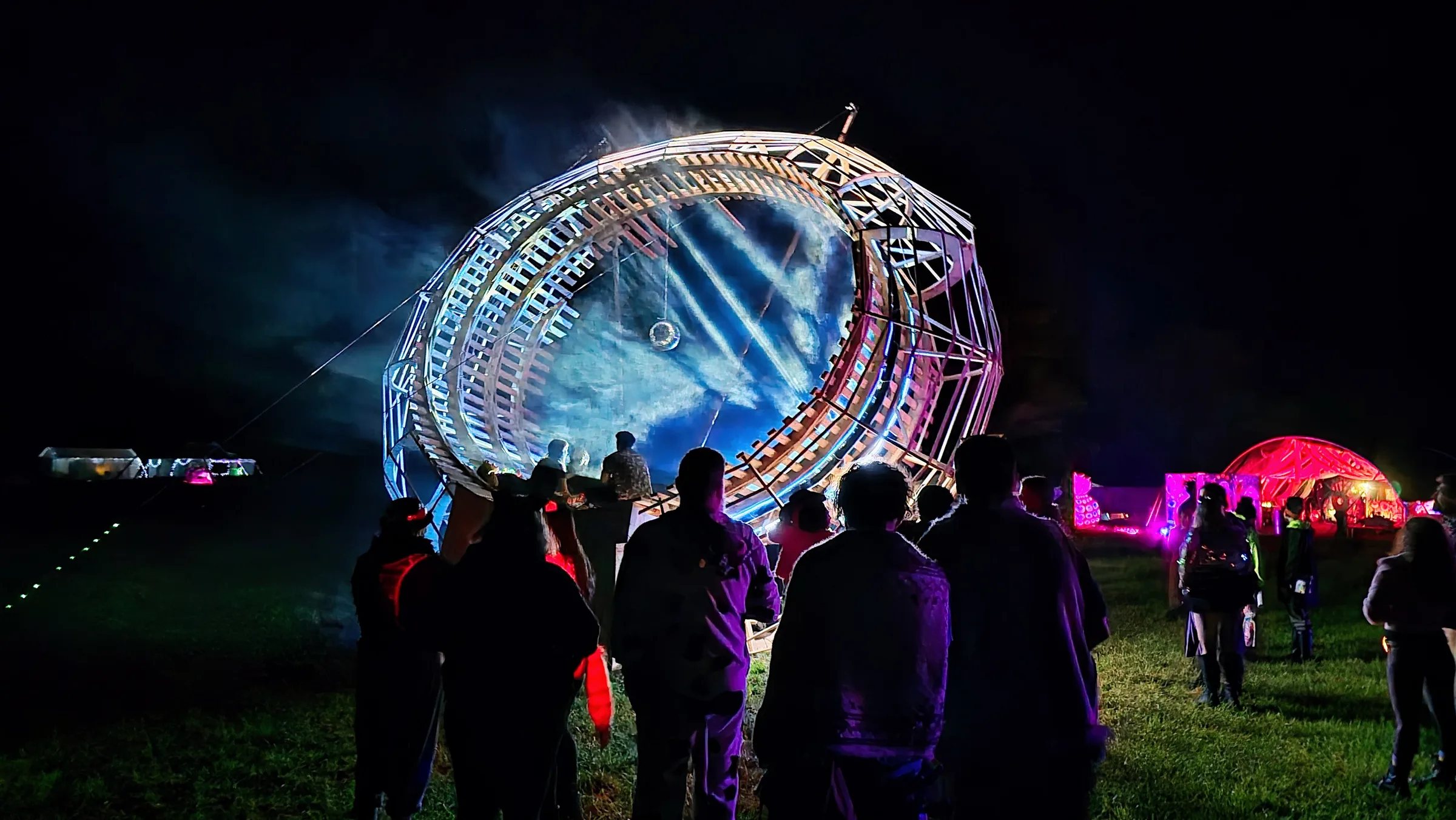 A crowd of silhouetted spectators faces the wooden ring glowing blue at night, a disco ball hanging inside and a pink tent glowing to the right.
