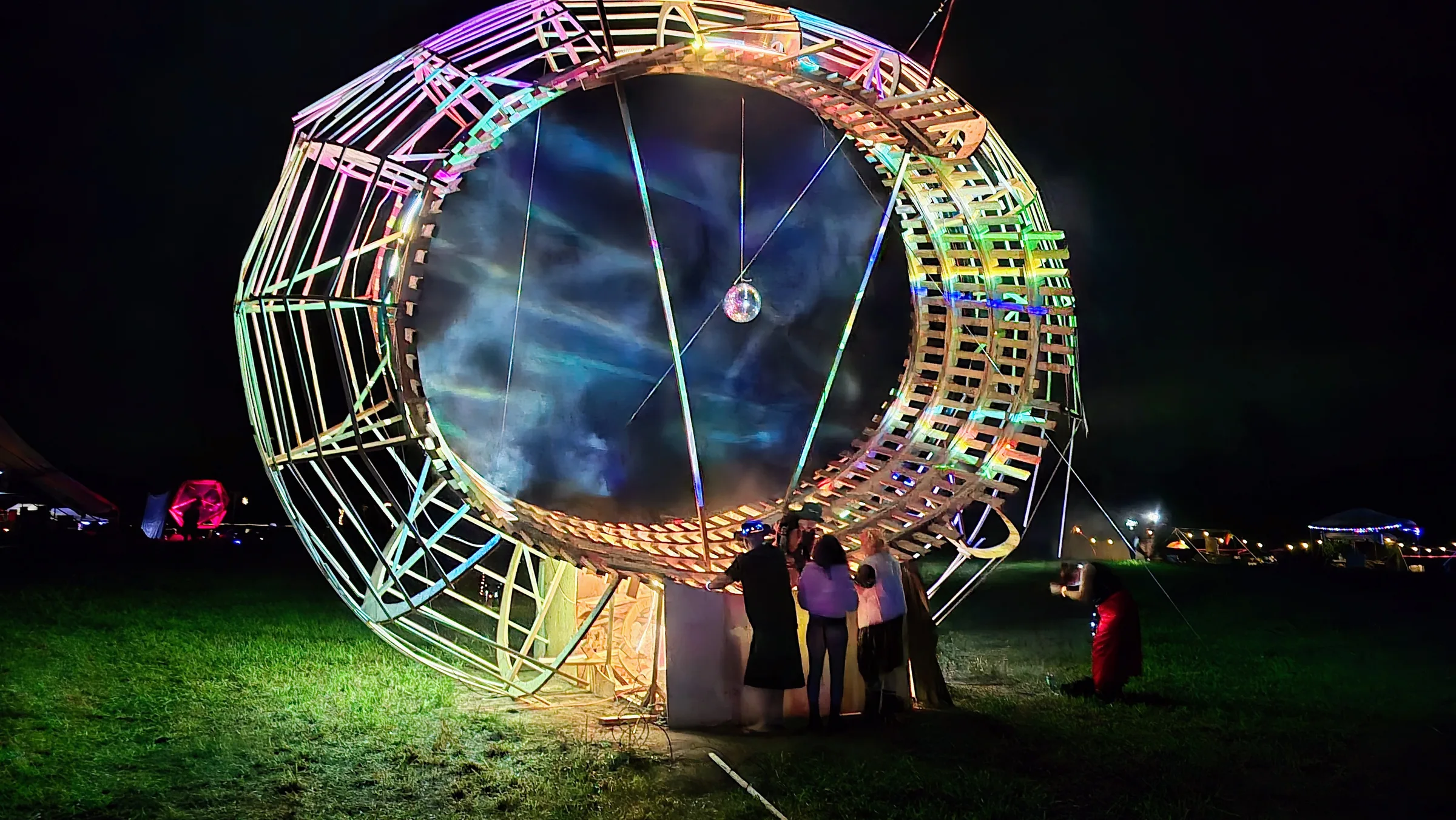 The wooden ring stands symmetrically at night with a dark, misty blue interior, a disco ball hanging center and small figures gathered at its base.