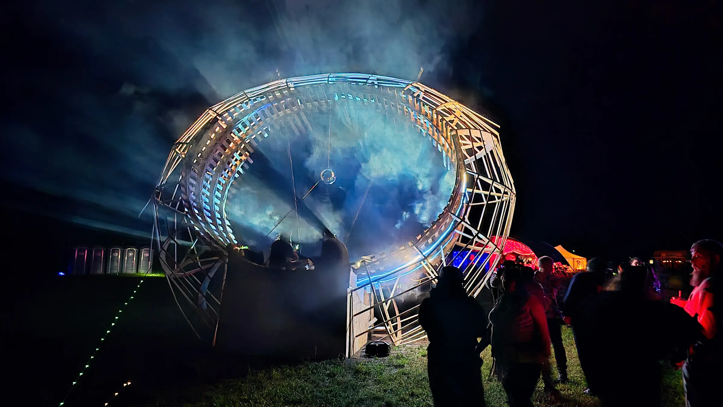 The illuminated ring radiates blue light beams into the night sky, silhouetted onlookers gathered nearby and colorful tents glowing in the distance.