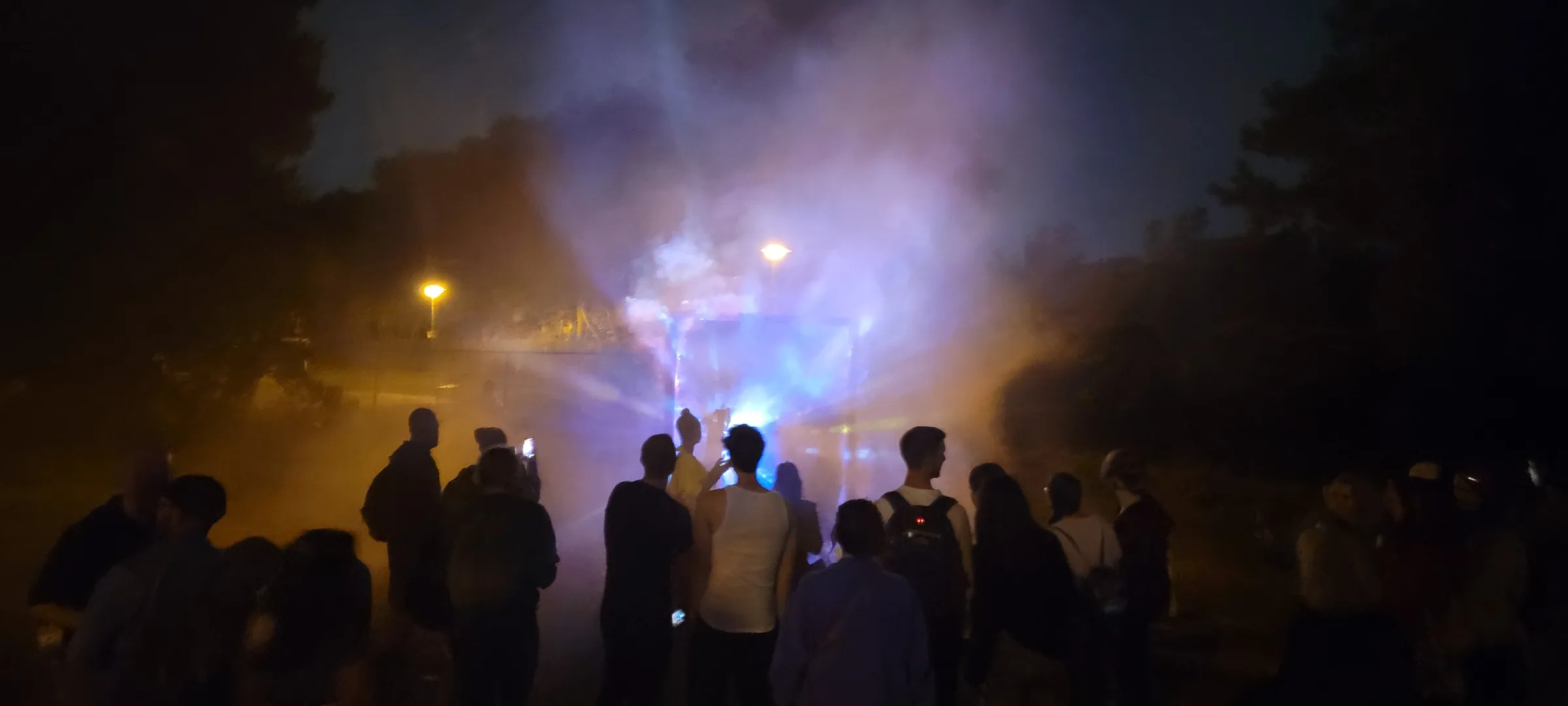Silhouetted onlookers at night watch a misty gateway glow purple and blue, some filming on phones, trees and streetlights in the background.