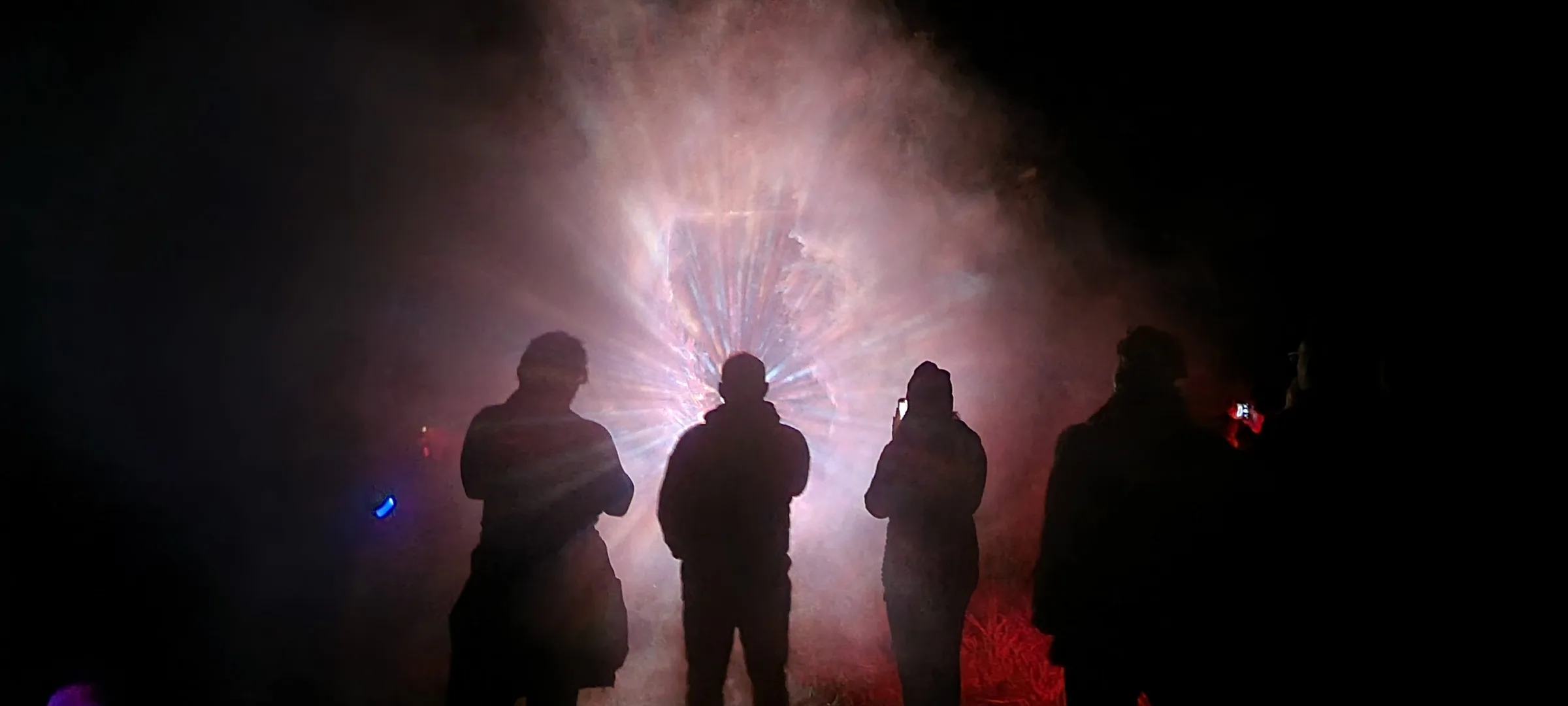Four silhouetted figures face a glowing rectangular portal at night, pink and white light rays bursting through fog, red light glowing on the ground.