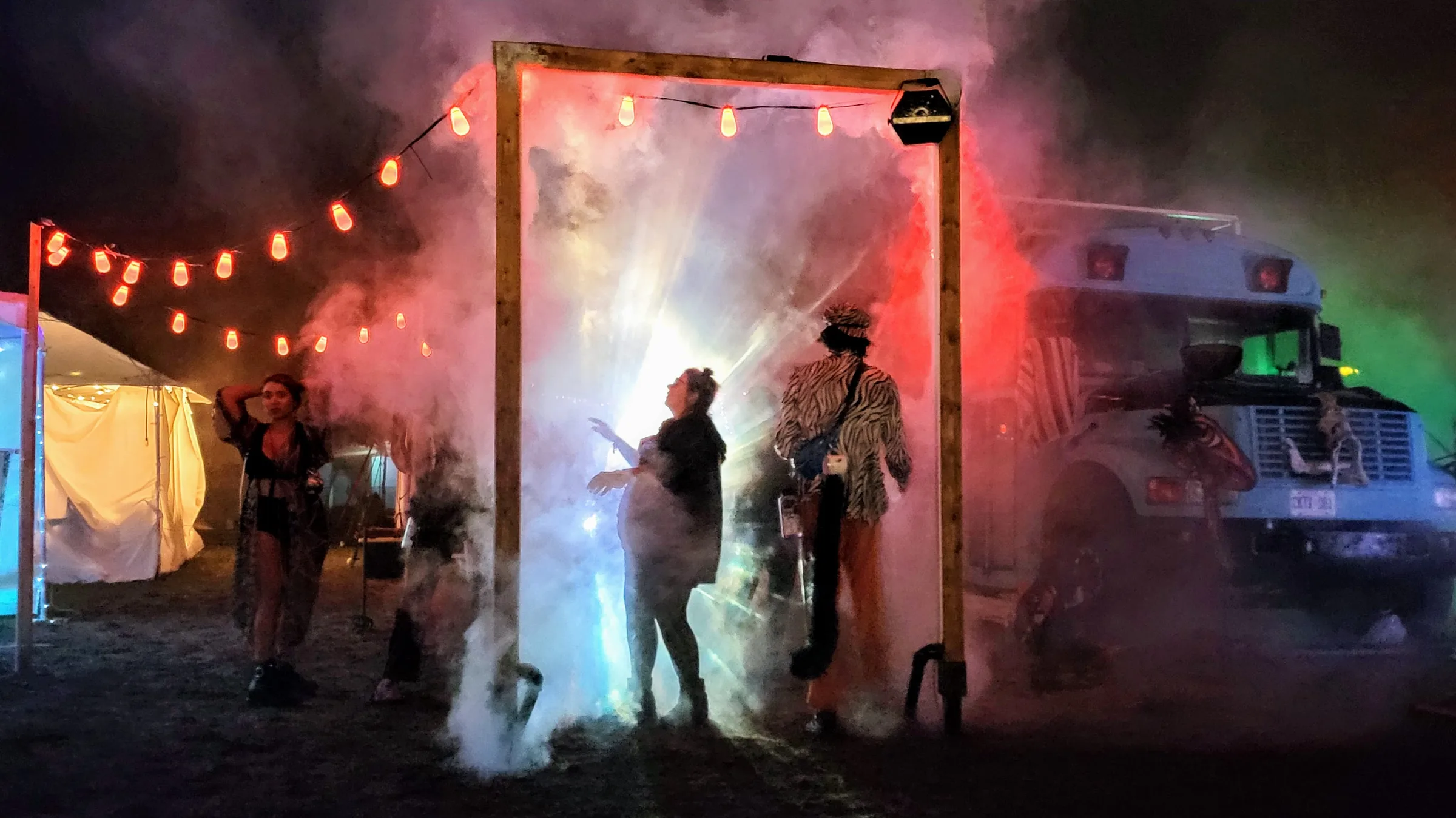 A wooden door frame strung with lights glows white through fog at a night festival, a silhouetted person inside, a bus and tents in the background.