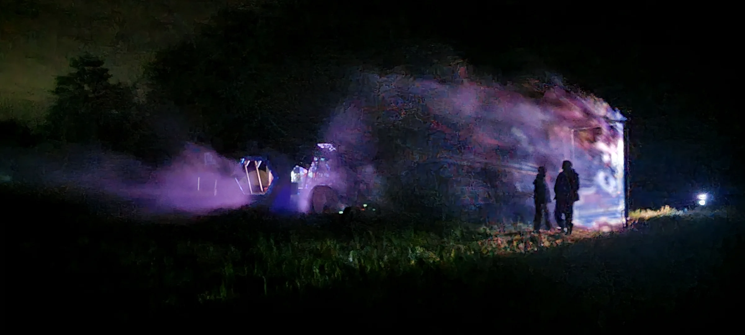 Two silhouetted people stand in a field at night, looking at a vehicle glowing with hazy purple projected light, trees dark against the sky.