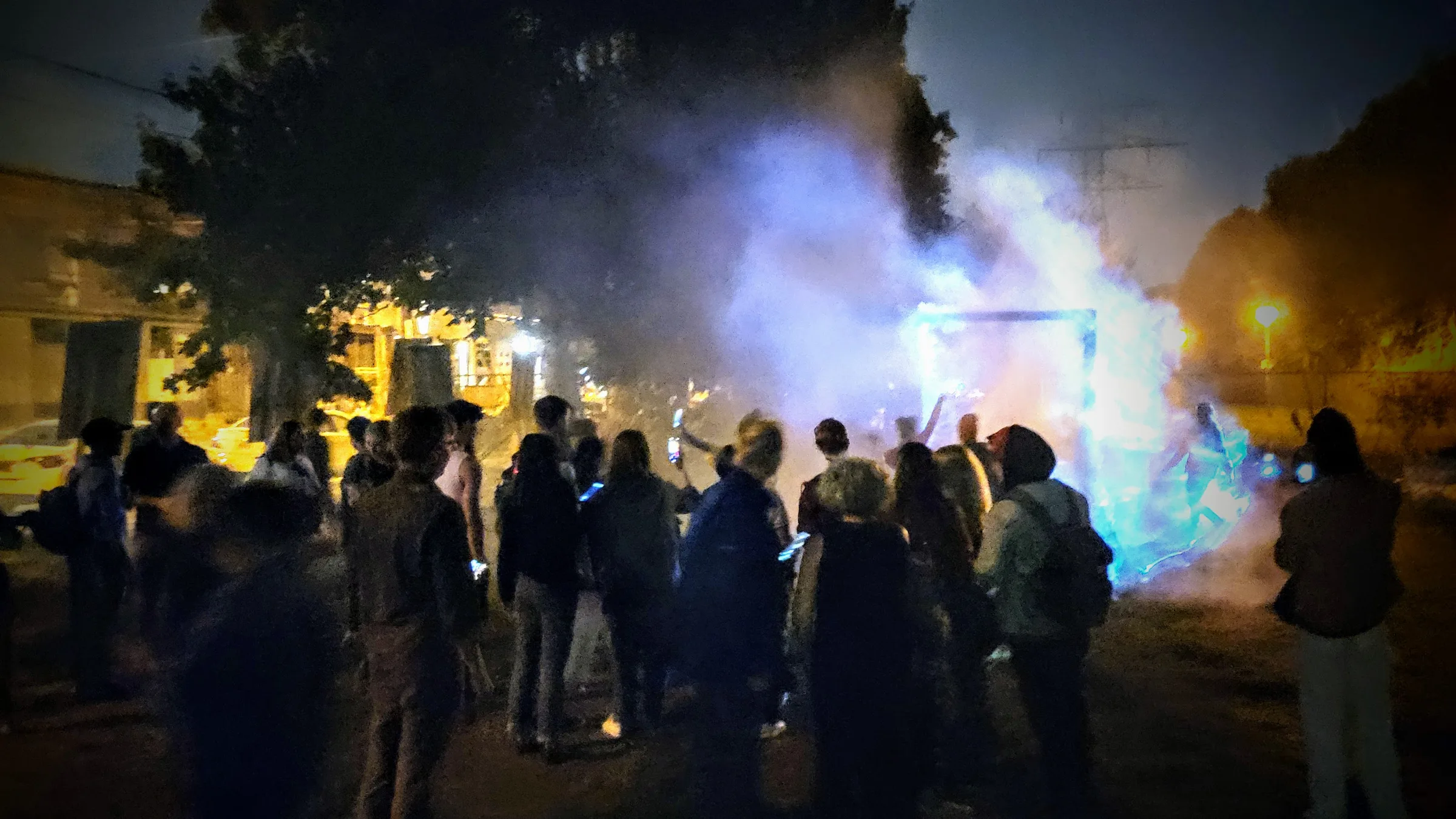 A dense nighttime crowd photographs a glowing blue portal frame billowing with lit fog, city buildings and a tree visible in the background.