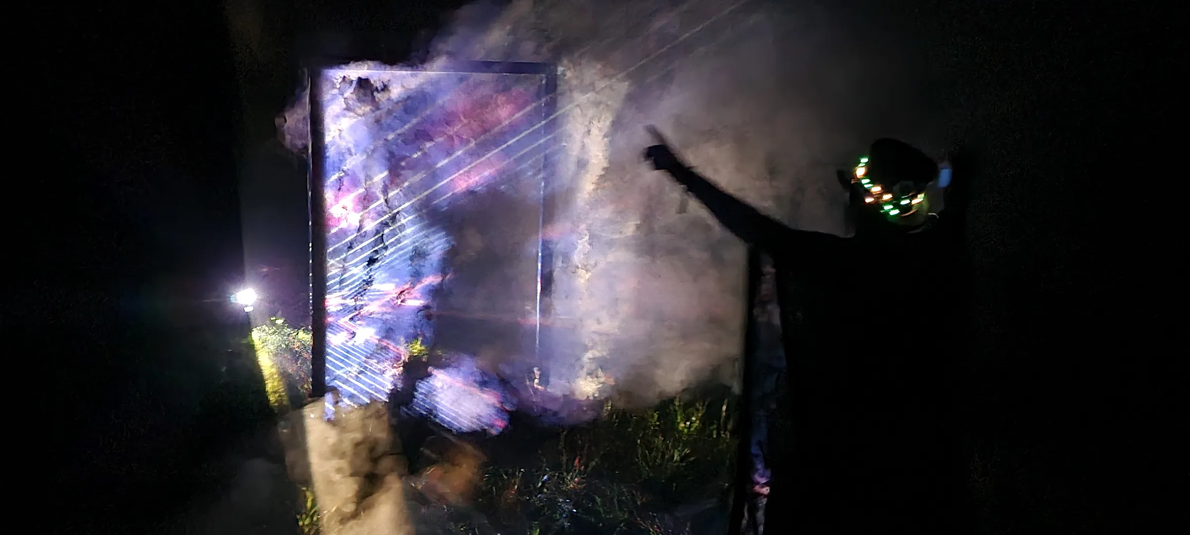 A cloaked figure wearing a glowing LED collar points toward a misty portal frame lit with crossing blue and pink light beams at night.