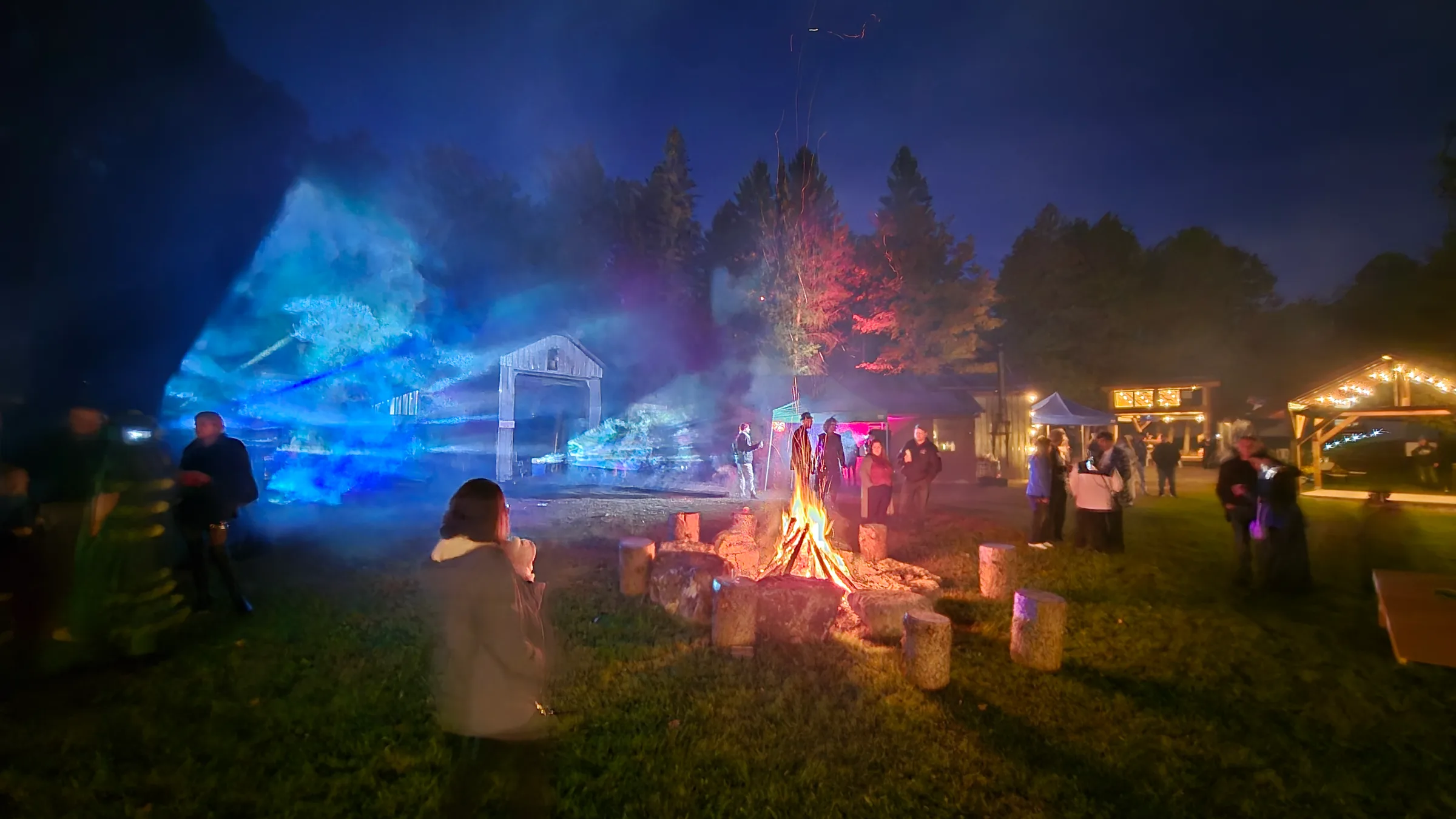 A nighttime crowd gathers on a lawn around a bonfire near a wooden barn, with blue-tinted light and haze glowing beside string-lit pavilions.